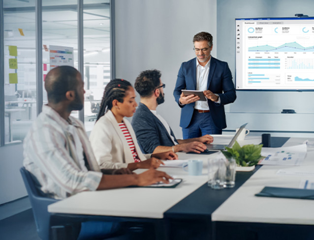 A business meeting with a diverse group of four people in an office. A man presents data on a screen, conveying professionalism and focus.