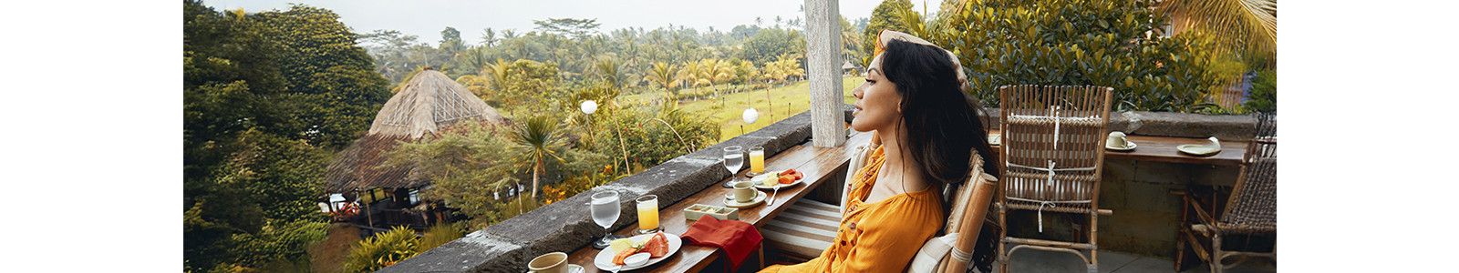 A woman on a balcony overlooks a verdant landscape, surrounded by trees and rolling hills under a clear sky.