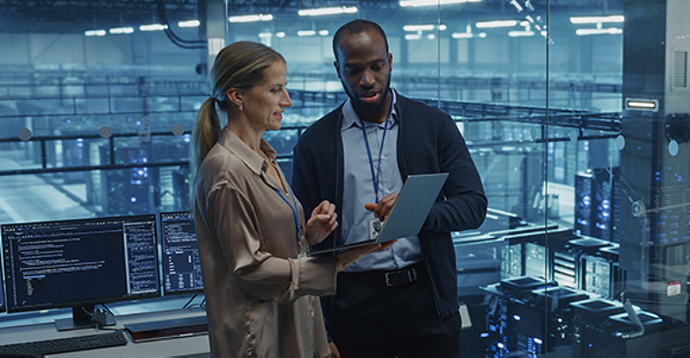Two professionals in a modern server room discuss data on a laptop. Monitors displaying code are visible, suggesting a tech-focused, collaborative environment.