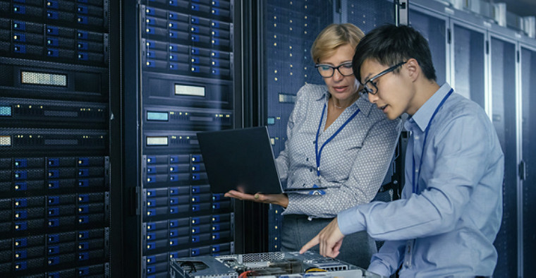 Two IT professionals in a server room, examining equipment. One holds a laptop, discussing data. The scene conveys focus and teamwork.