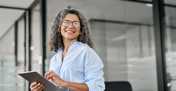 A smiling woman with curly gray hair and glasses holds a tablet in a modern office. She wears a blue-striped shirt, conveying confidence and ease.