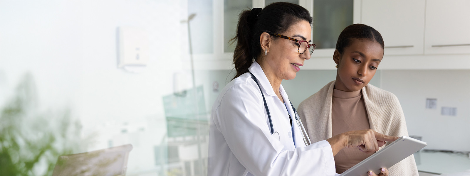 A woman wearing a white coat is focused on a tablet, appearing engaged with the content displayed on the screen.
