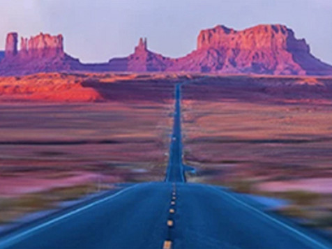 A long stretch of highway road with red rocky mountains in the background.