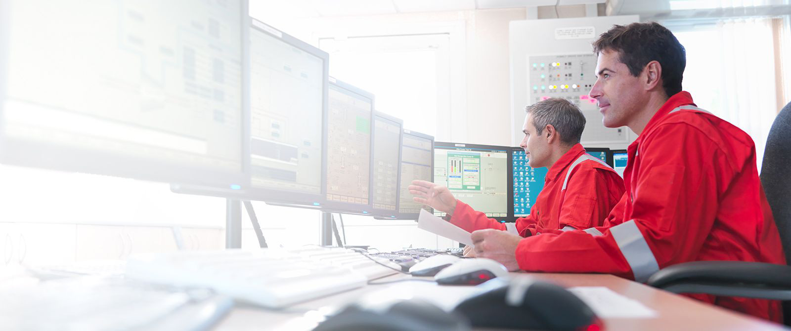 Two men in high-visibility safety suits monitoring something on their desktop computers