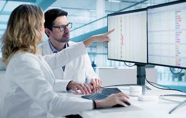 Two scientists in lab coats focus on dual monitors displaying data. A woman points at the screen, discussing with a colleague, in a modern lab setting.