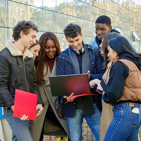 Un grupo diverso de hombres y mujeres jóvenes de pie alrededor de un hombre y mirando un laptop