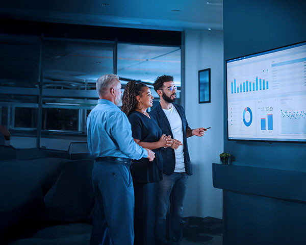 Three individuals stand in front of a large screen, engaged in discussion.
