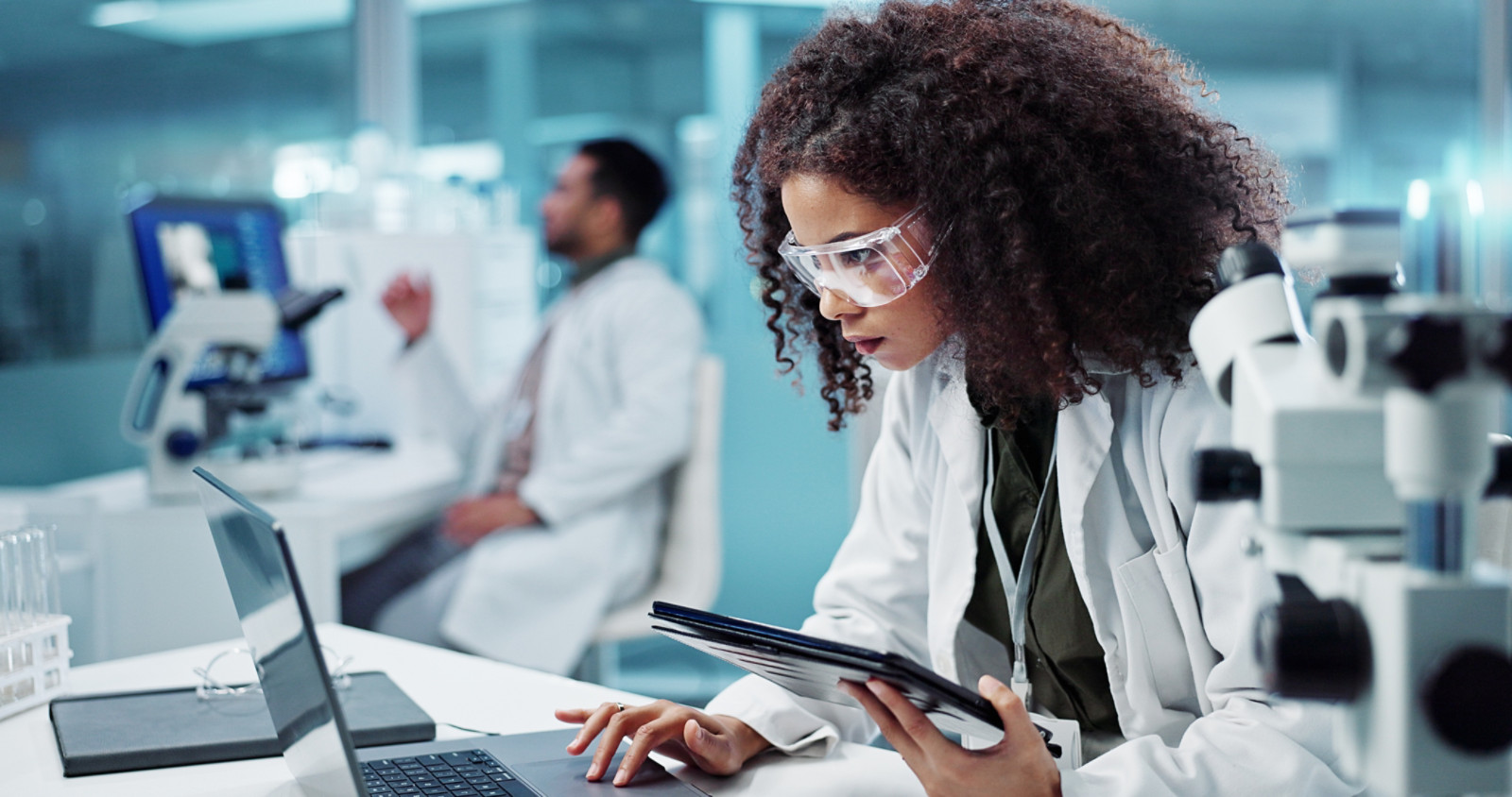 A lab technician wearing protective glasses, holding a tablet in one hand and working on a laptop with the other