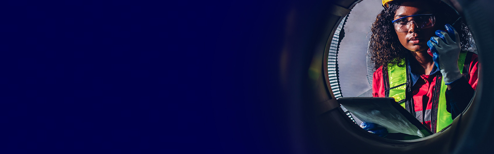 A woman wearing a hazmat jacket, safety glasses and gloves, holding a clipboard and a walkie-talkie while looking out of a porthole