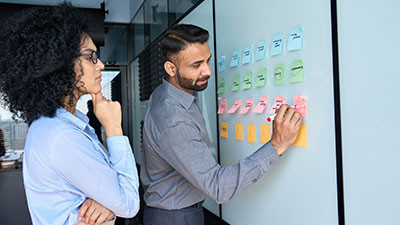 A man and a woman standing in front of a whiteboard consisting of several sticky notes, on which the man is writing something
