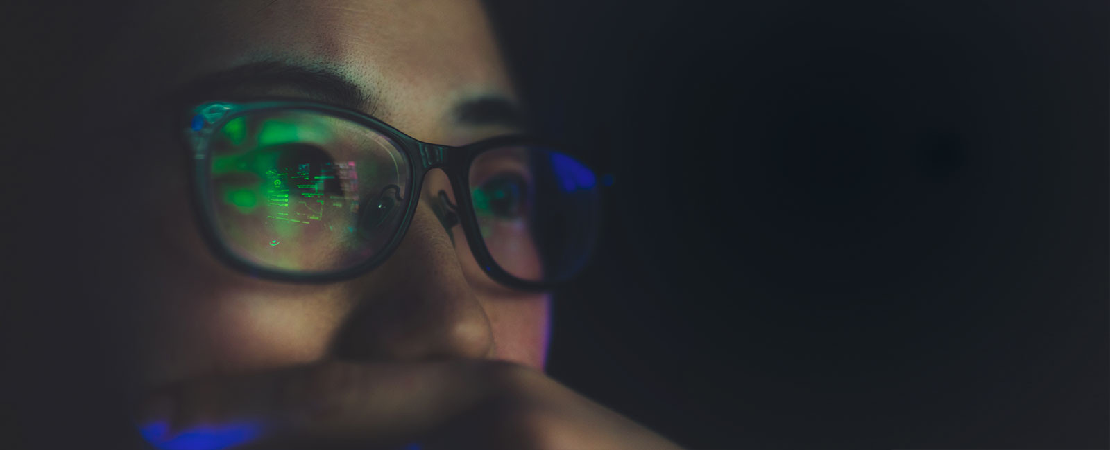 A closeup view of a bespectacled woman, with the screen she is looking at being reflected on her glasses