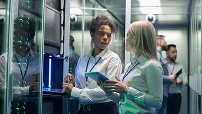 In a server room, two female co-workers talking and further behind them are two male co-workers looking at the servers