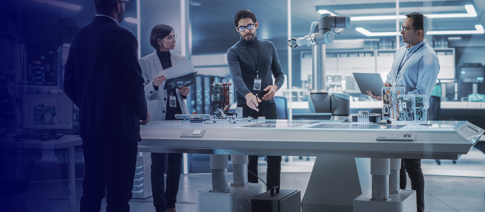 Three men and a woman are standing around an electronic display table, while one of the men is explaining something