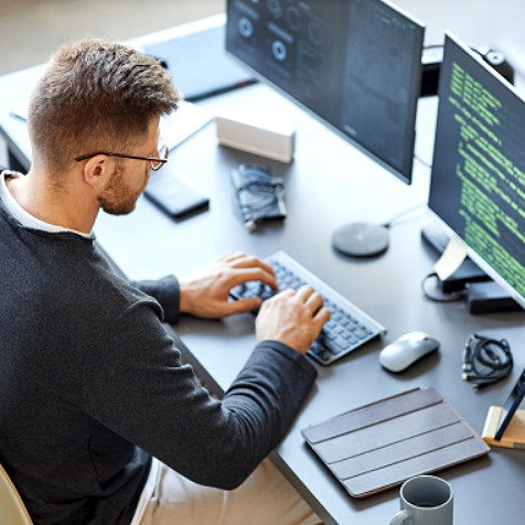 Young man sitting at his desk typing on his keyboard and looking at two computer monitors