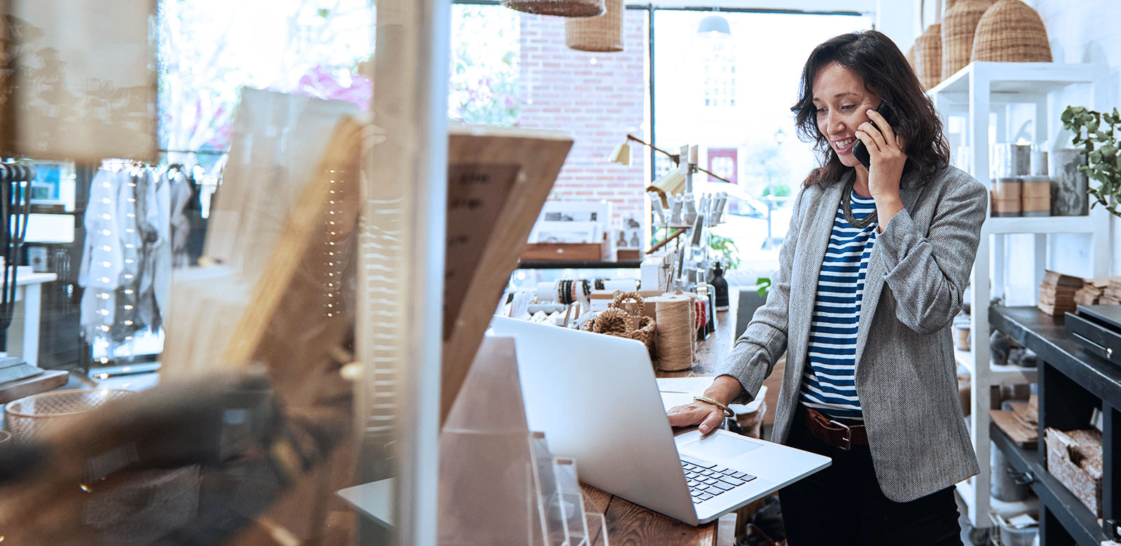 A woman is on the phone while working on her laptop in a lively shop environment