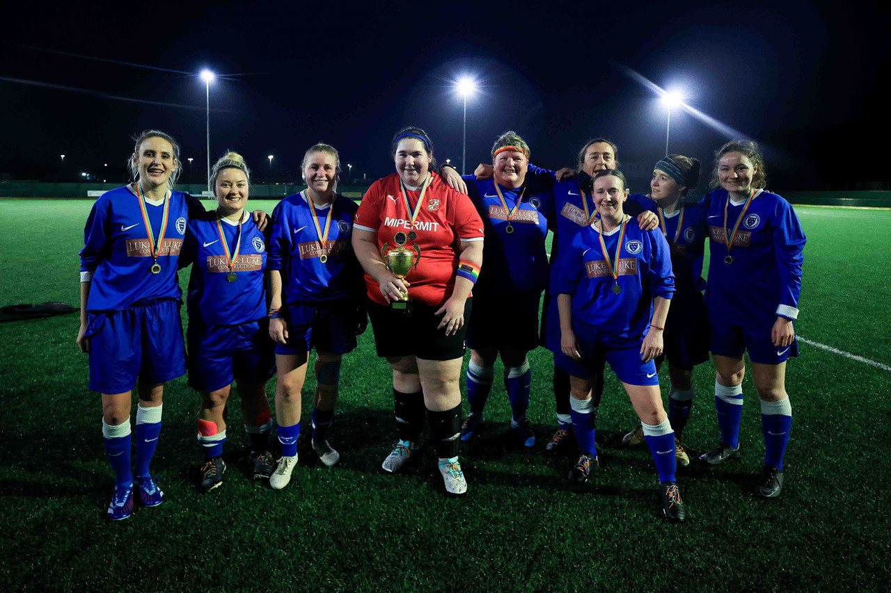 elley Tiltman stands with her teammates, representing the Sussex FA IAG.  They have just won the Football vs Homophobia charity match (February 2024). The team is wearing a blue football kit and are smiling, holding medals and a trophy. 