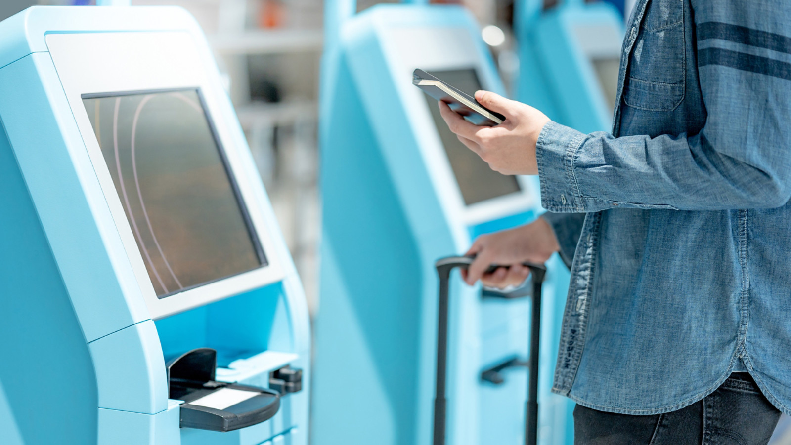 Tourist holding passport and smartphone using self check-in kiosk in airport terminal