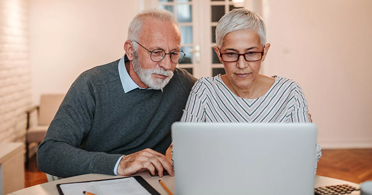 Two older adults sitting side by side at a table, looking at and using a laptop computer in a bright indoor setting.