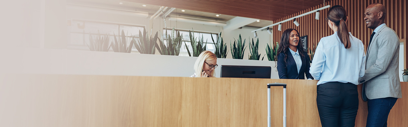 A hotel receptionist standing and conversing with a couple, while a second customer care executive sits smiling and working on her computer.